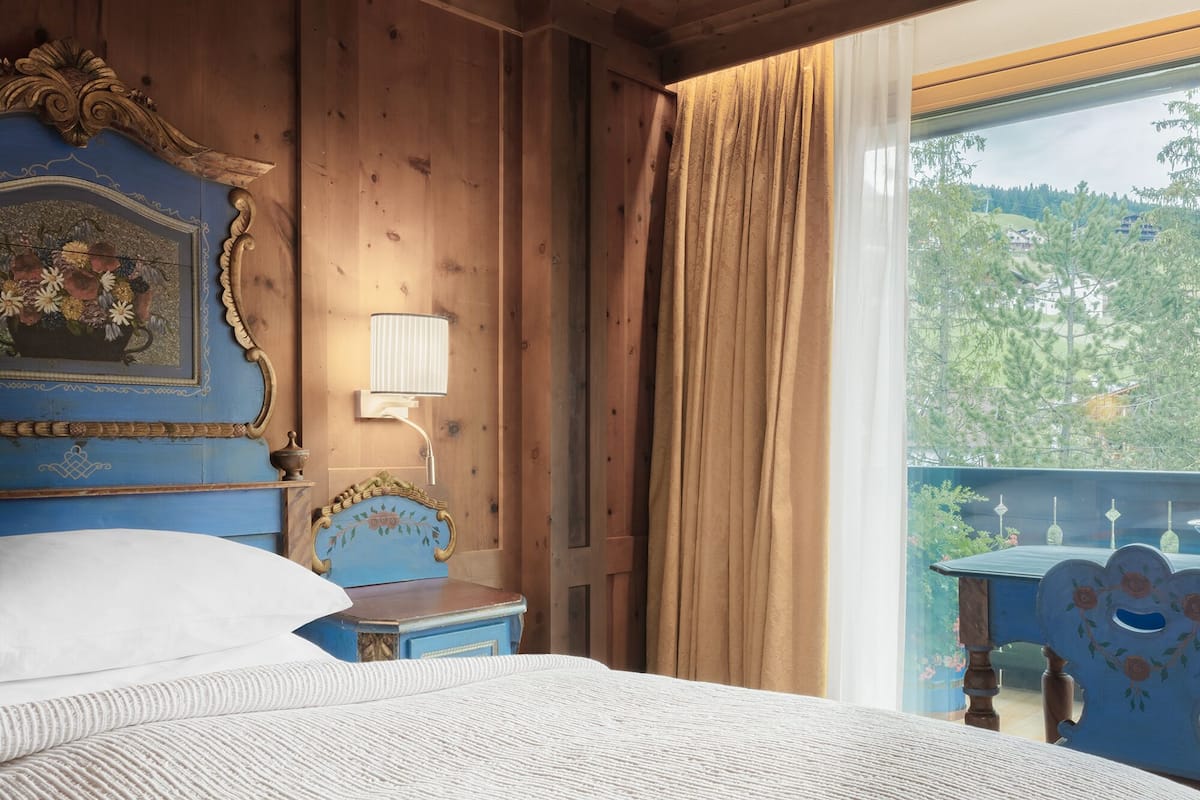 Een slaapkamer met een houten hoofdeinde, een bed met wit beddengoed, een nachtkastje, een wandlamp en een raam met gordijnen.