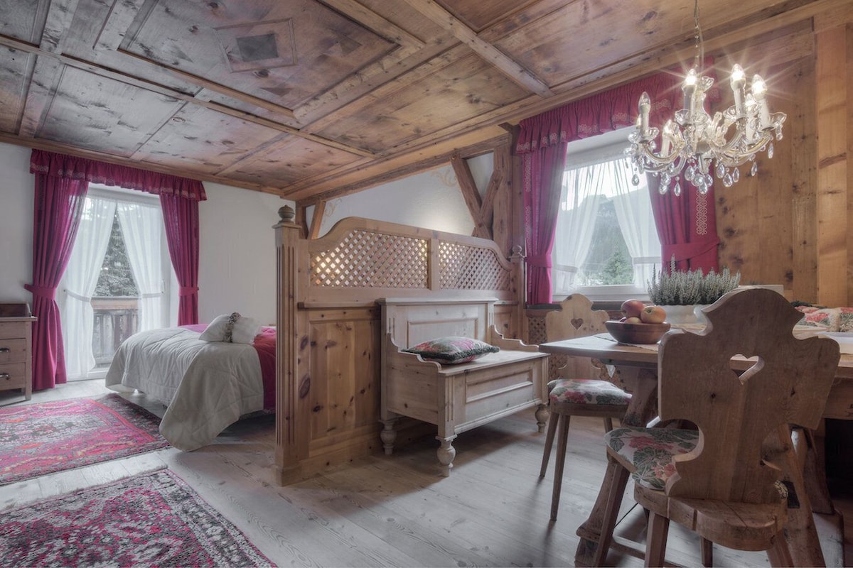 A cozy bedroom with a wooden bed, a desk, and a chair, a chandelier, and a view of the outdoors.
