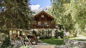 A rustic wooden cabin with a stone foundation, surrounded by lush greenery and a clear mountain view.
