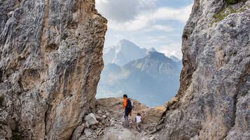 Sendero montañoso pedregoso con una persona y un niño caminando por una estrecha abertura entre acantilados, con vistas a montañas distantes.