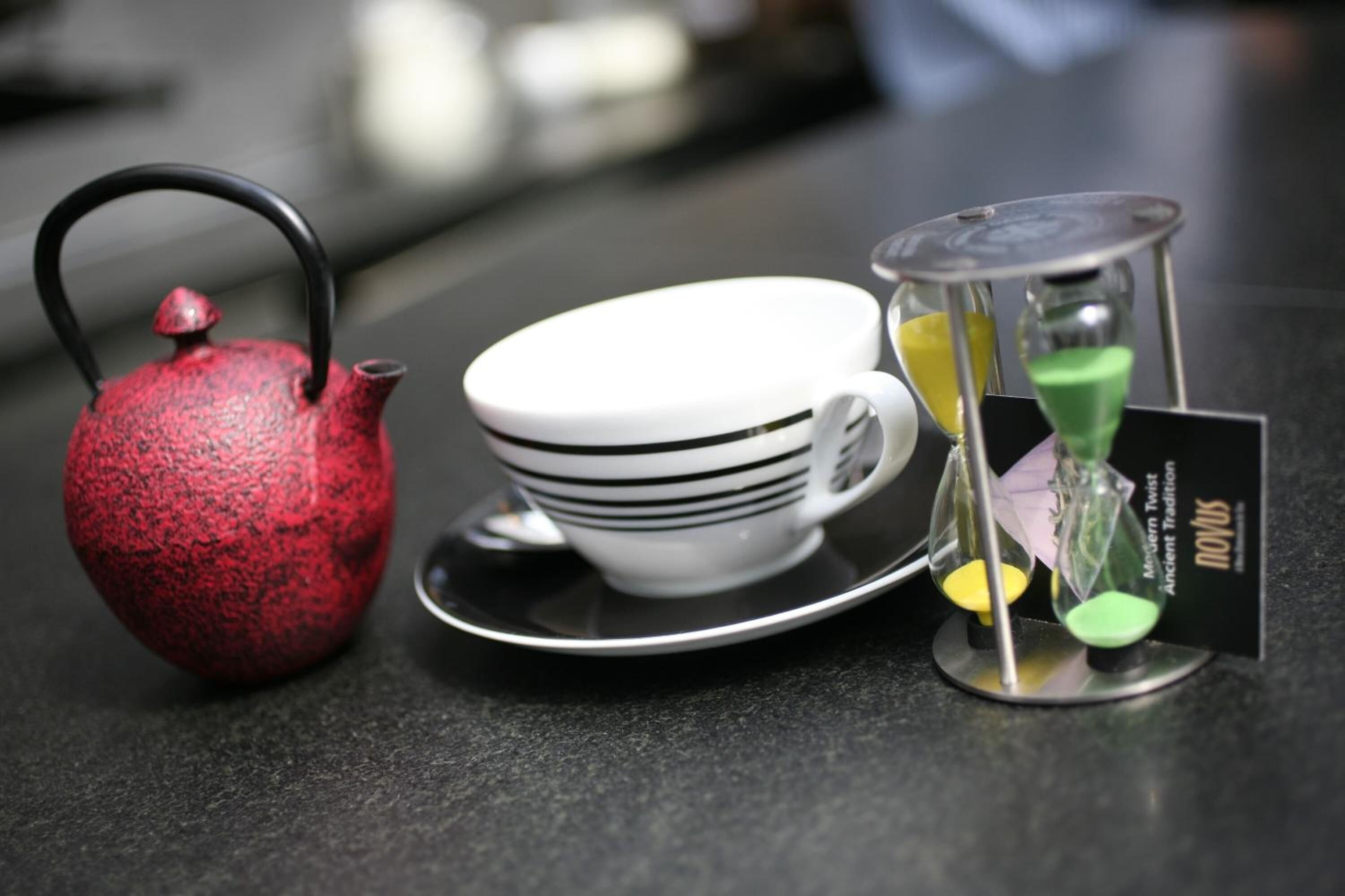 A red teapot, a white cup with black stripes, and a small hourglass on a black surface.
