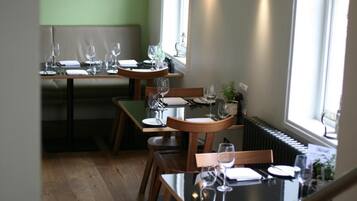 Un área de comedor con mesas y sillas de madera, una pared verde con un aplique en forma de flor, y una ventana que proporciona luz natural.