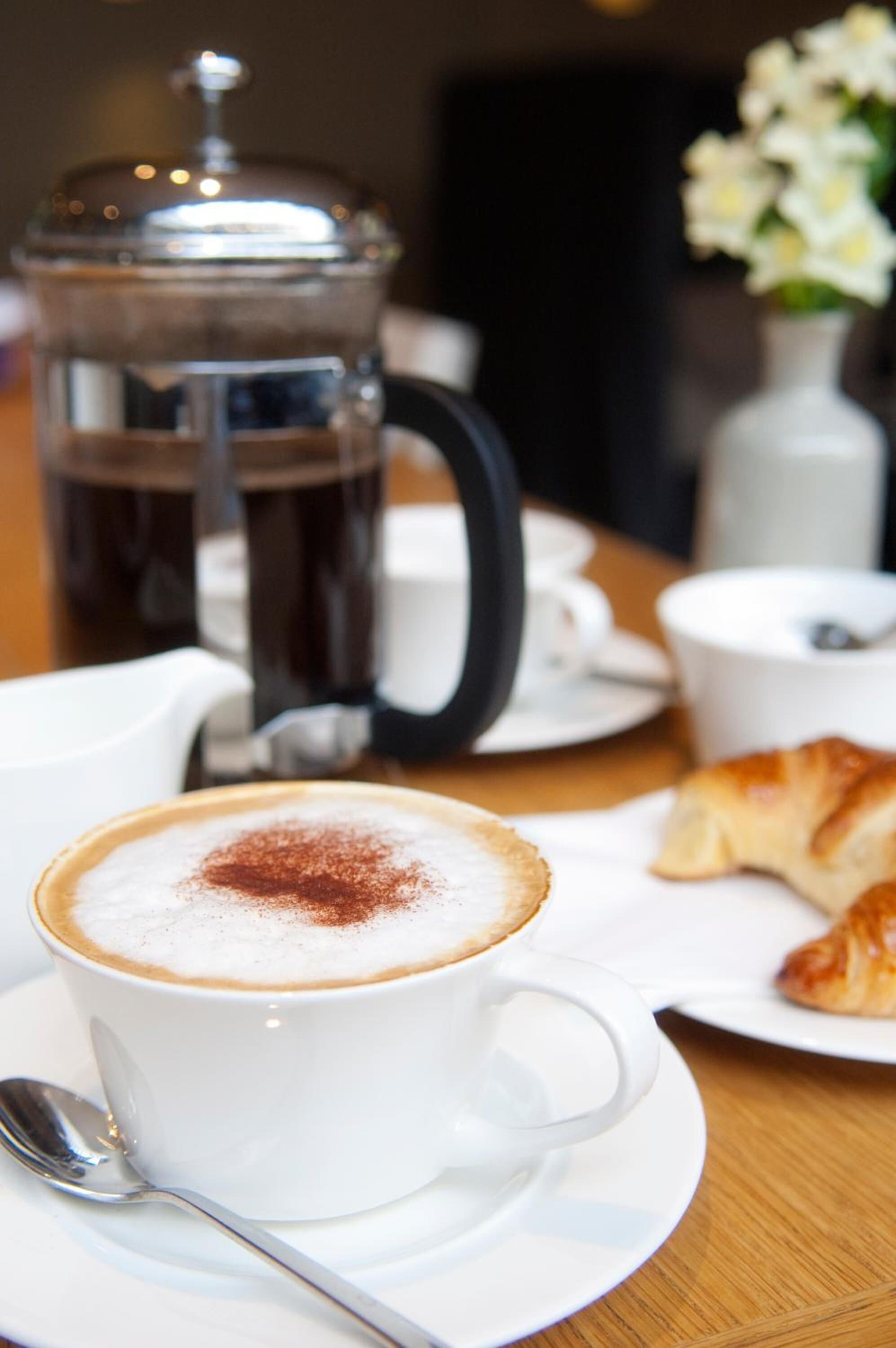 A cup of coffee with a frothy top and a sprinkle of cinnamon on a saucer, a croissant, a coffee pot, and a small white cup with a spoon on a wooden table.