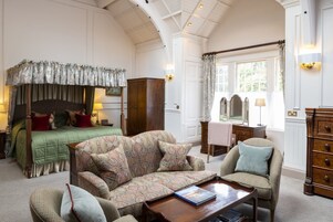 Um quarto espaçoso com cama de dossel, um sofá, uma poltrona, cômoda de madeira e uma escrivaninha com abajur.