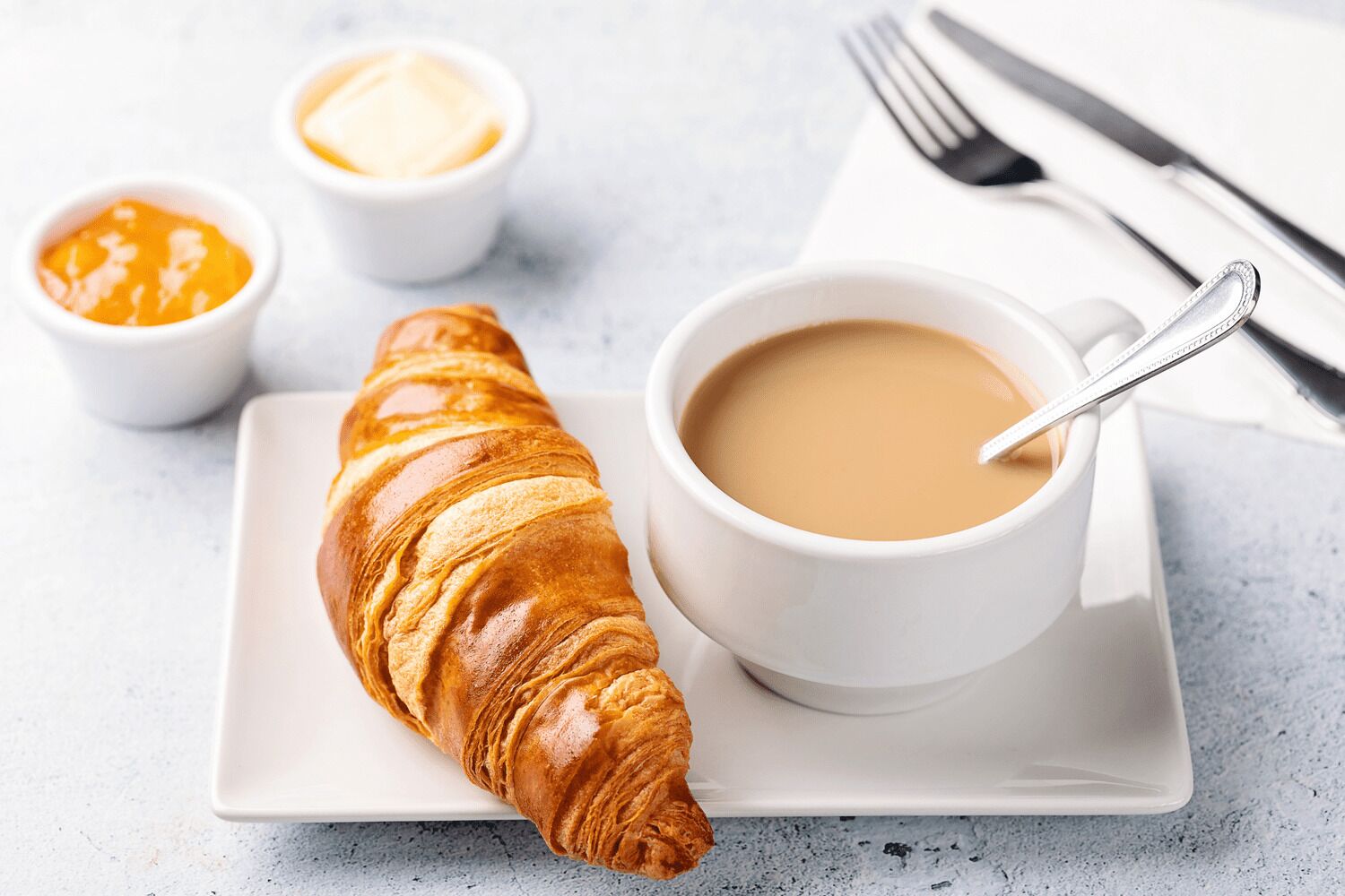 Et croissanter på en hvid tallerken, en kop kaffe med en ske, og to små skåle med smør.