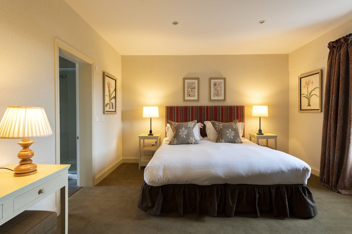 A hotel room with a large bed, two bedside tables with lamps, a desk with a lamp, and a door leading to a shower.