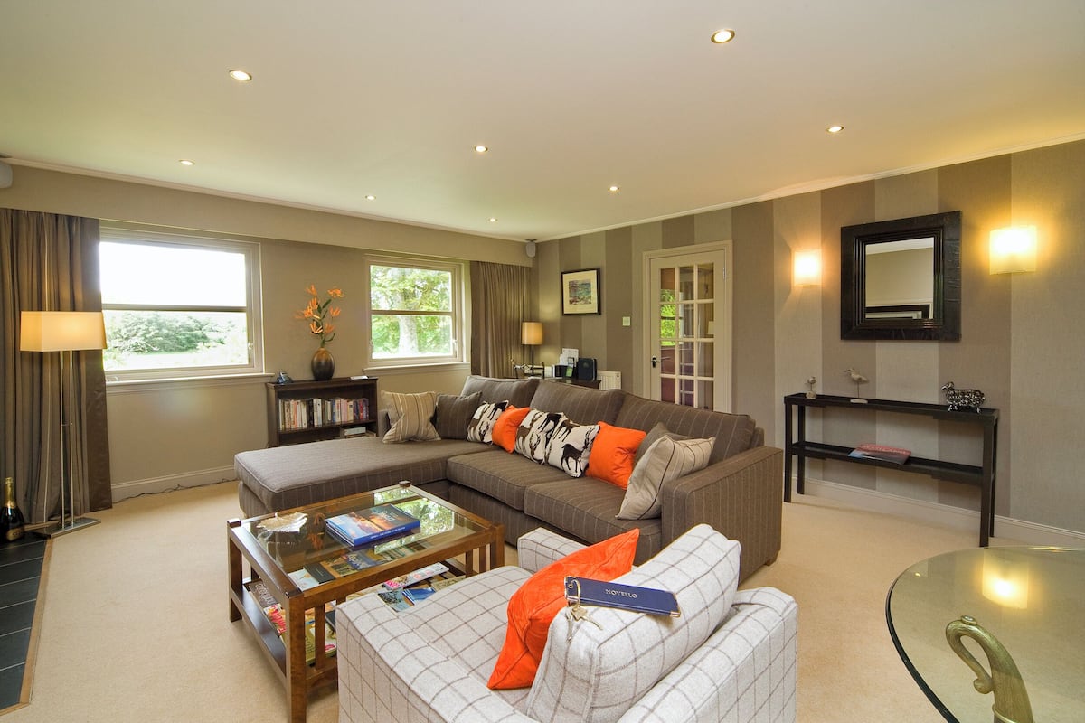 A living room with a sectional sofa, a glass coffee table, a bookshelf, and a mirror on the wall.
