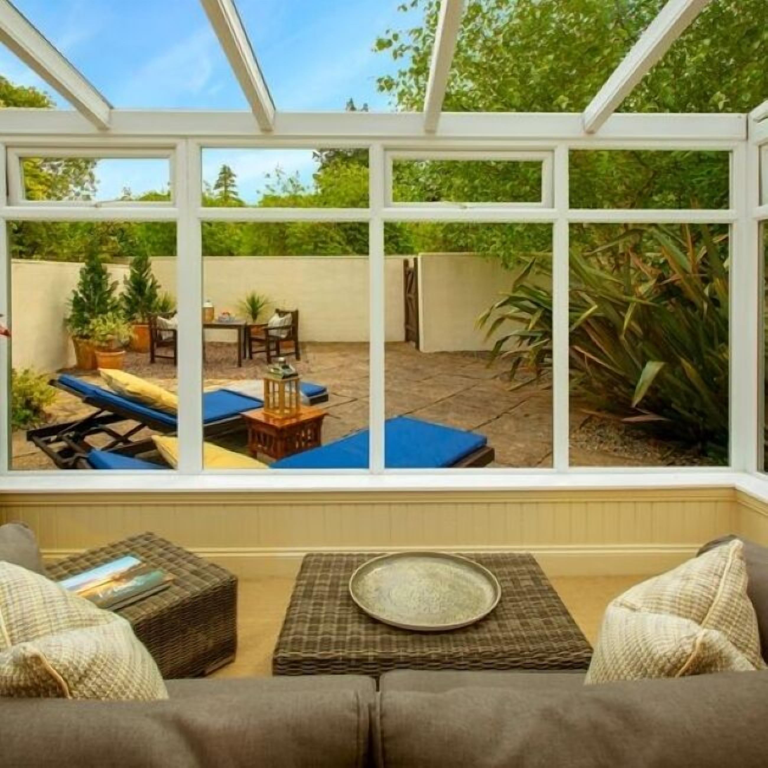 A conservatory with a view of a patio area featuring outdoor furniture, a table, and potted plants.