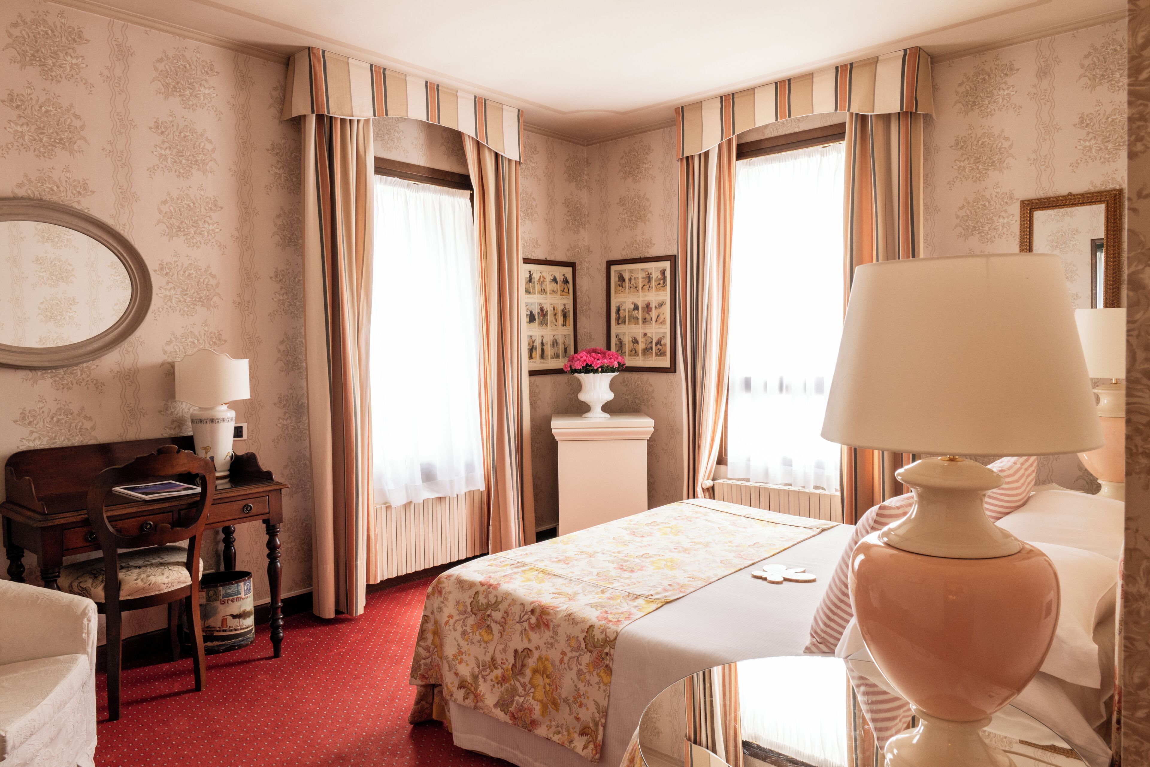 A hotel room with a bed, a desk, a chair, a lamp, a radiator, and a wall with framed pictures.