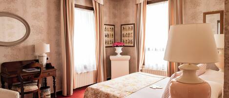 A hotel room with a bed, a desk, a chair, a lamp, a radiator, and a wall with framed pictures.