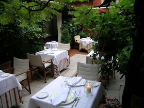 Un área de comedor al aire libre con mesas dispuestas para dos personas, rodeada de abundante follaje.