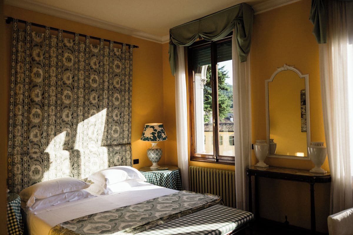 Habitación de hotel con cama, una mesa con lámpara, un espejo y una ventana con cortinas.