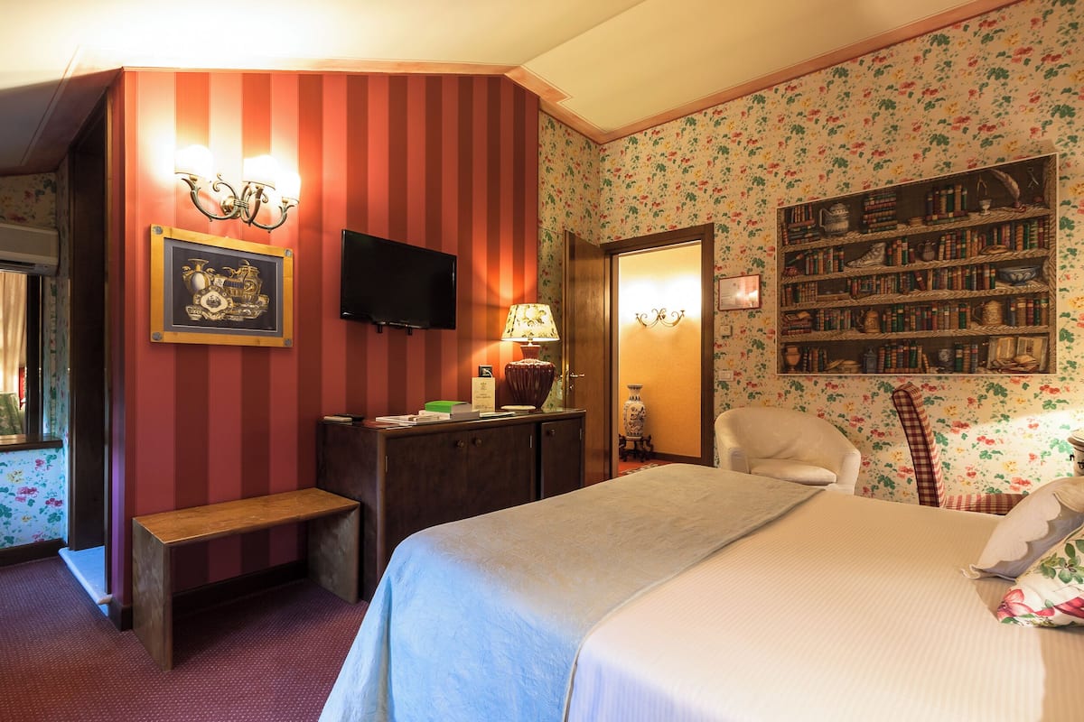 Una habitación de hotel con una cama, un banco, un televisor, una estantería y un estampado de flores en el papel tapiz.