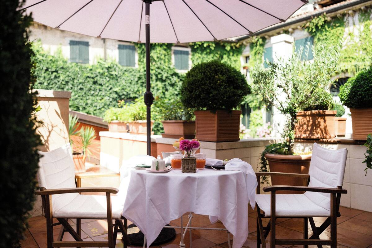 An outdoor patio with a table set for two, surrounded by white cushioned chairs, under a large umbrella.