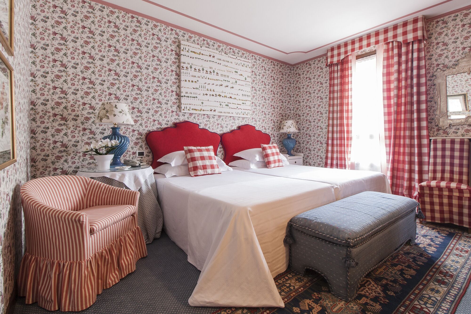 Une chambre à coucher avec un papier peint à motifs floraux, un lit double avec une literie rouge et blanche, un fauteuil à rayures et un pouf à carreaux.