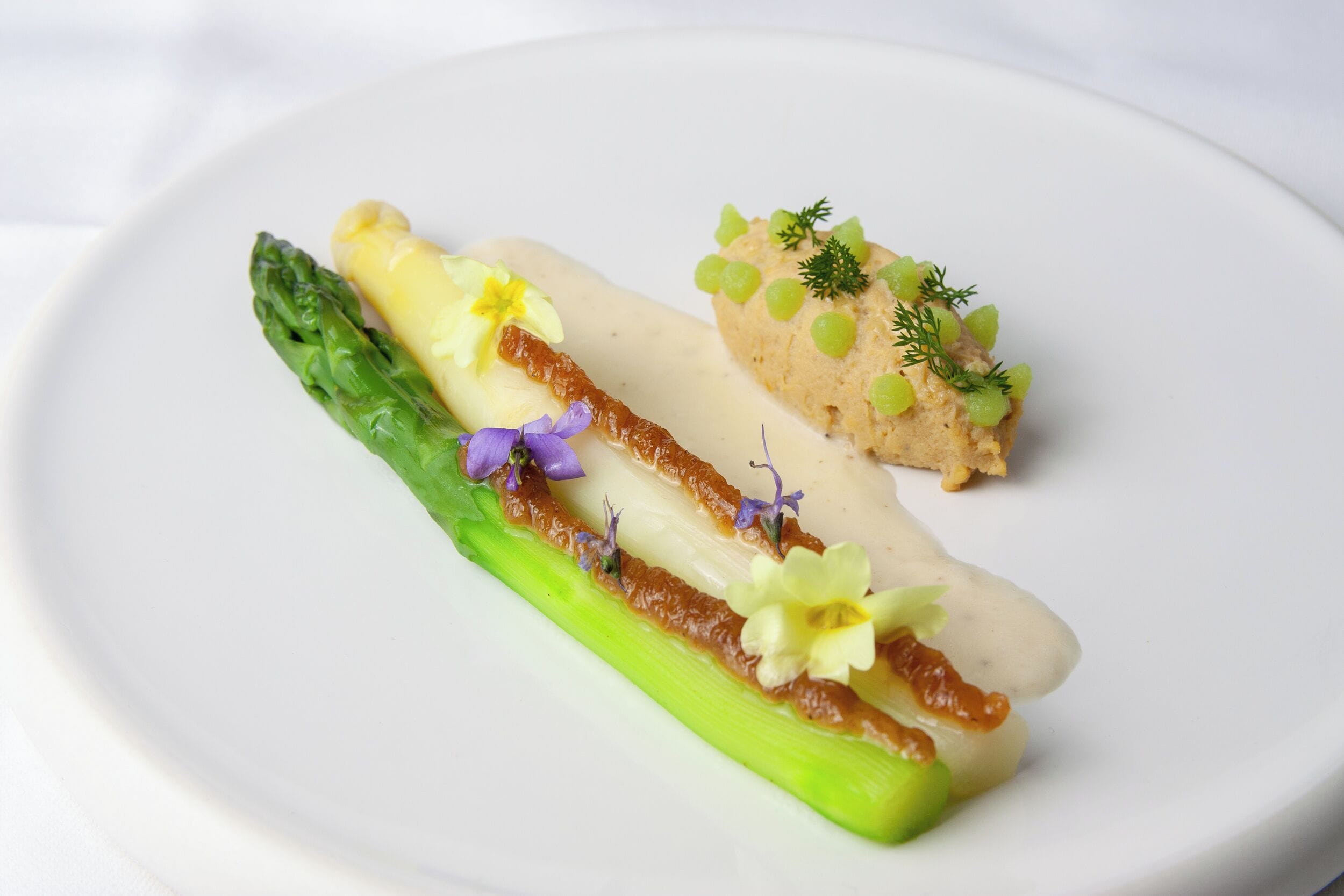 A white plate with a dish of food, including asparagus and a creamy substance, garnished with green and purple elements.