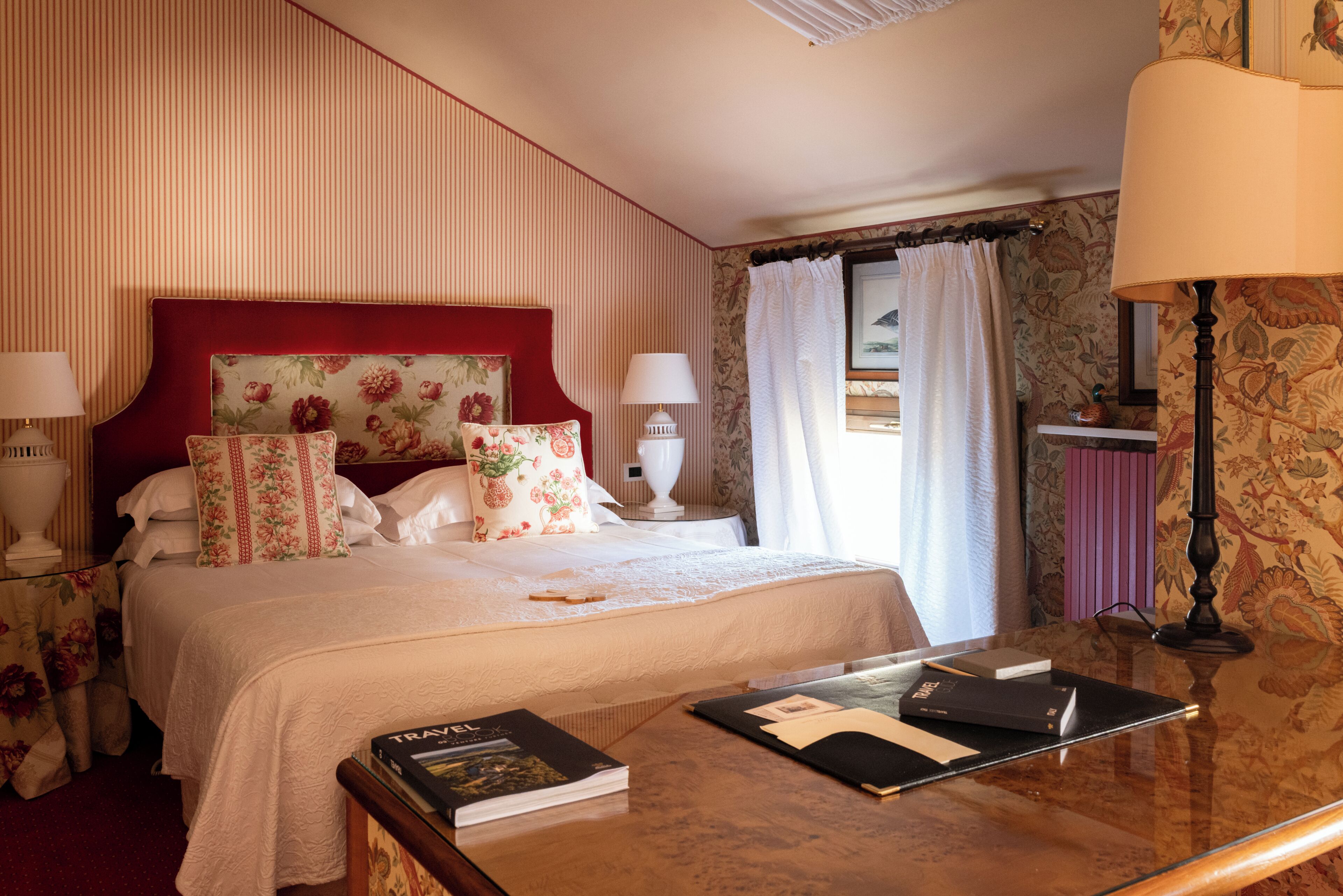 A hotel room with a large bed, a bedside table with a lamp, a window with curtains, and a wooden nightstand.