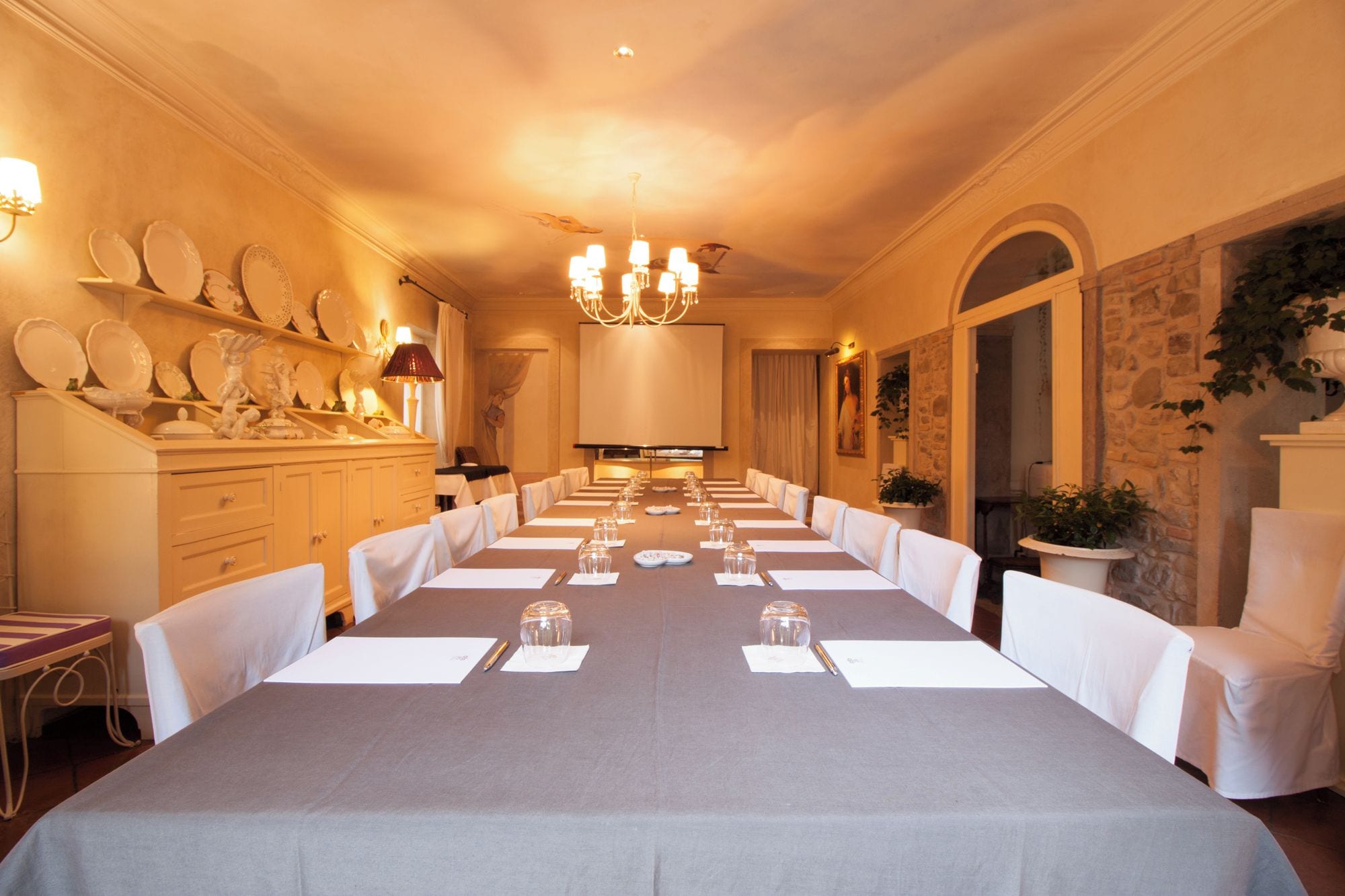 Una larga mesa de comedor dispuesta para un evento formal, con sillas, una araña y una pared de piedra con plantas.