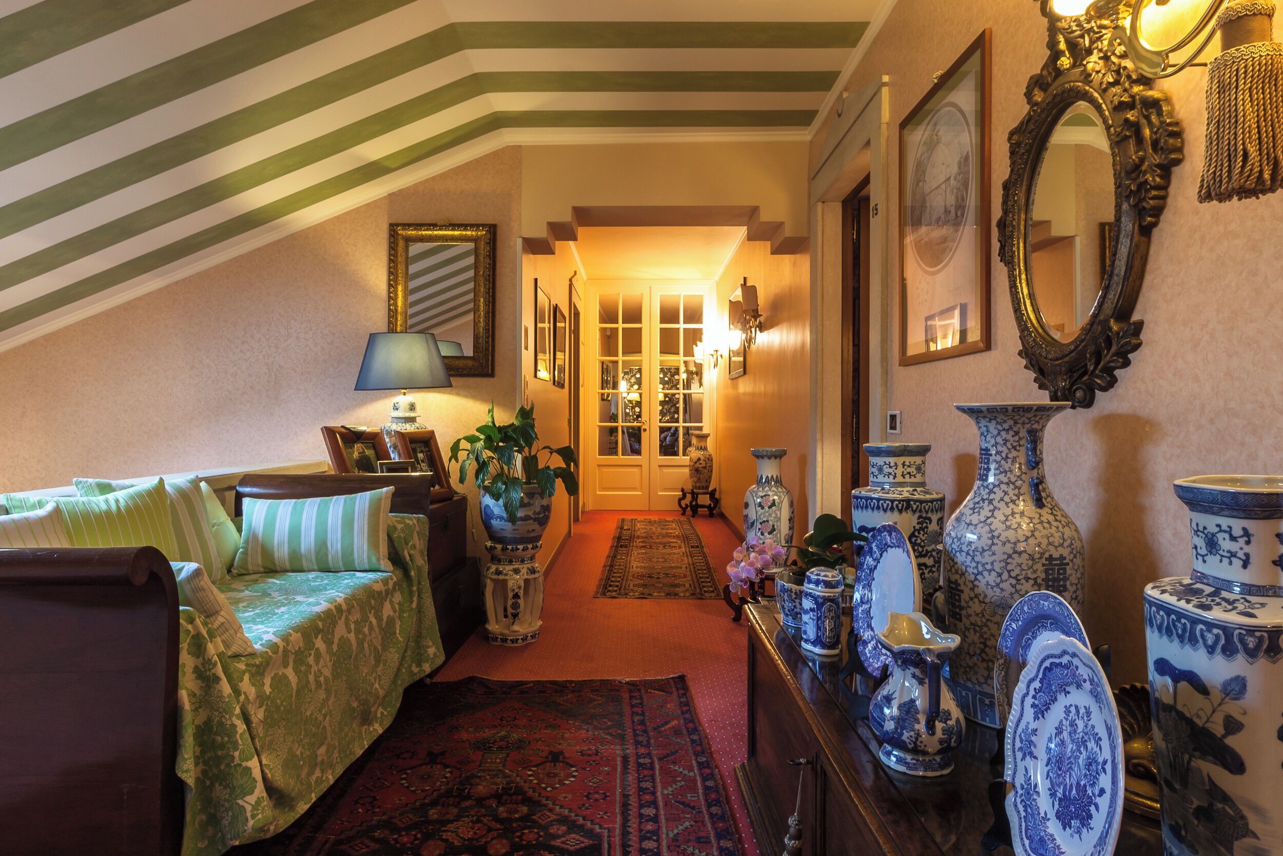 A cozy room with a sloped ceiling, a bed with green bedding, a wooden dresser with blue and white porcelain, and a hallway leading to a door.