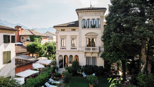 A courtyard with a large building featuring arched windows and a balcony, surrounded by lush greenery and outdoor seating. - Relais & Chateaux Villa Abbazia (Follina)