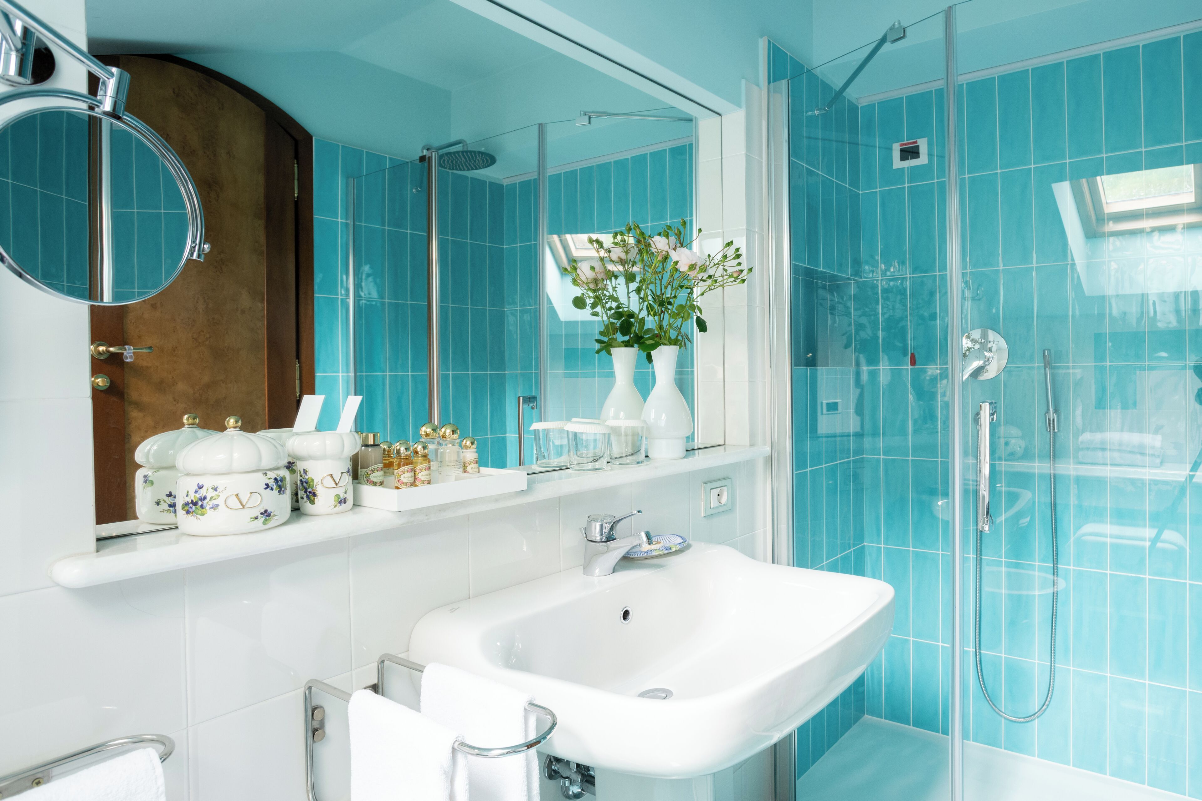 A bathroom with a white sink, a mirror, a shower, and a vase with flowers on the wall.