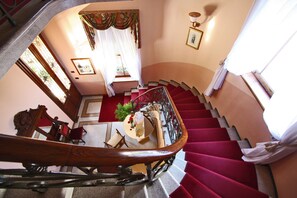 A grand staircase with red carpeted steps and a wooden handrail, leading to a room with a round table, chairs, and a small plant. - Relais & Chateaux Villa Abbazia (Follina)