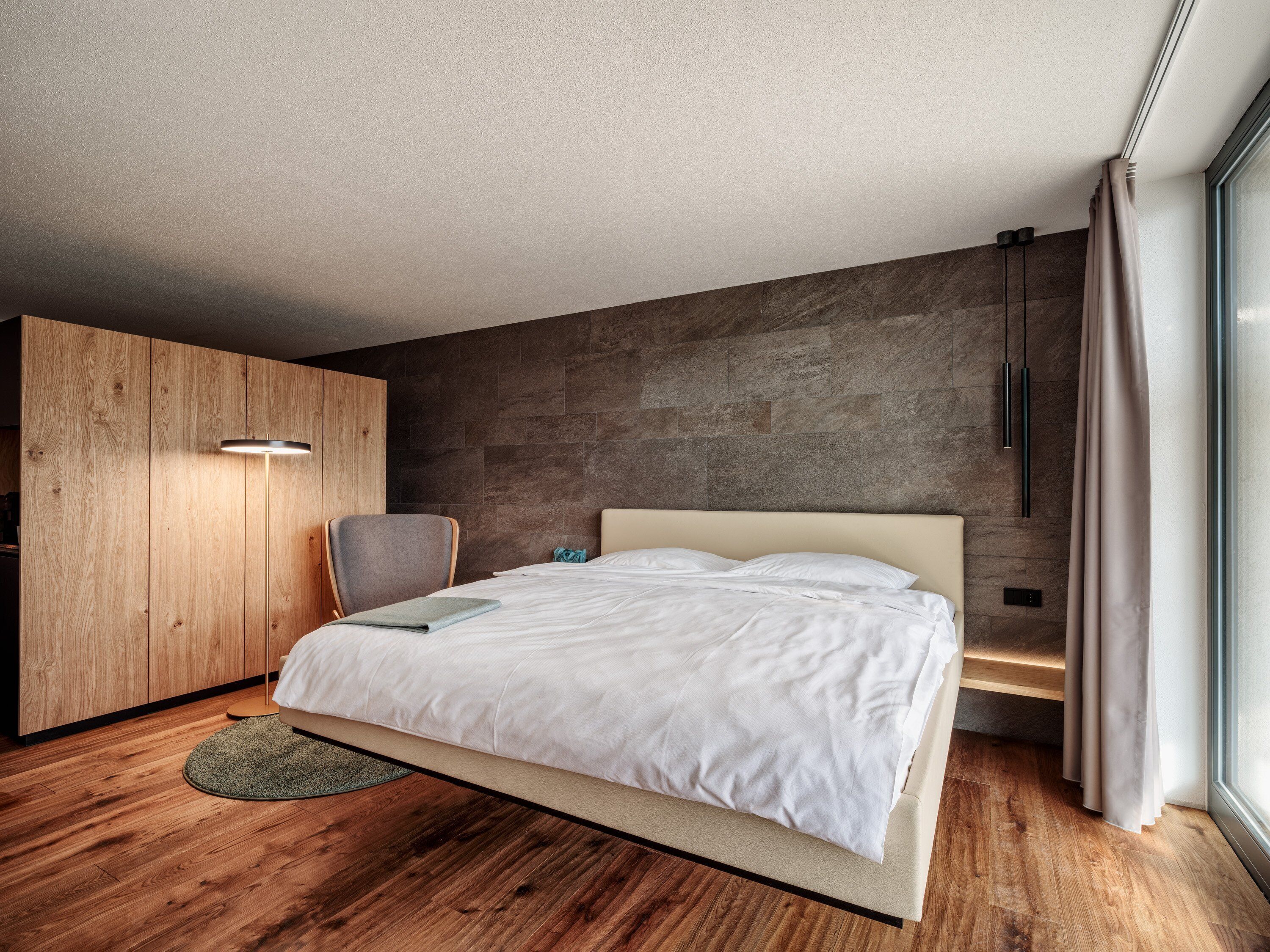 A modern bedroom with a wooden wardrobe, a bed with white bedding, a grey chair, and a stone wall.