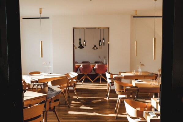 Comedor con mesas y sillas de madera, iluminación colgante y vista a un espacio con sillas rojas.