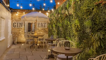 Enjoy meals in a charming outdoor dining area with a stone wall, round tables, and lush greenery.