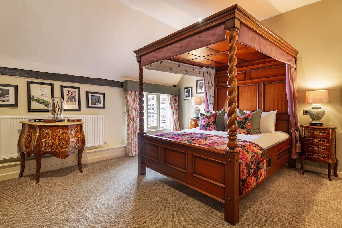 A four-poster bed with a floral bedspread, a wooden nightstand with drawers, and a table with a floral tablecloth.