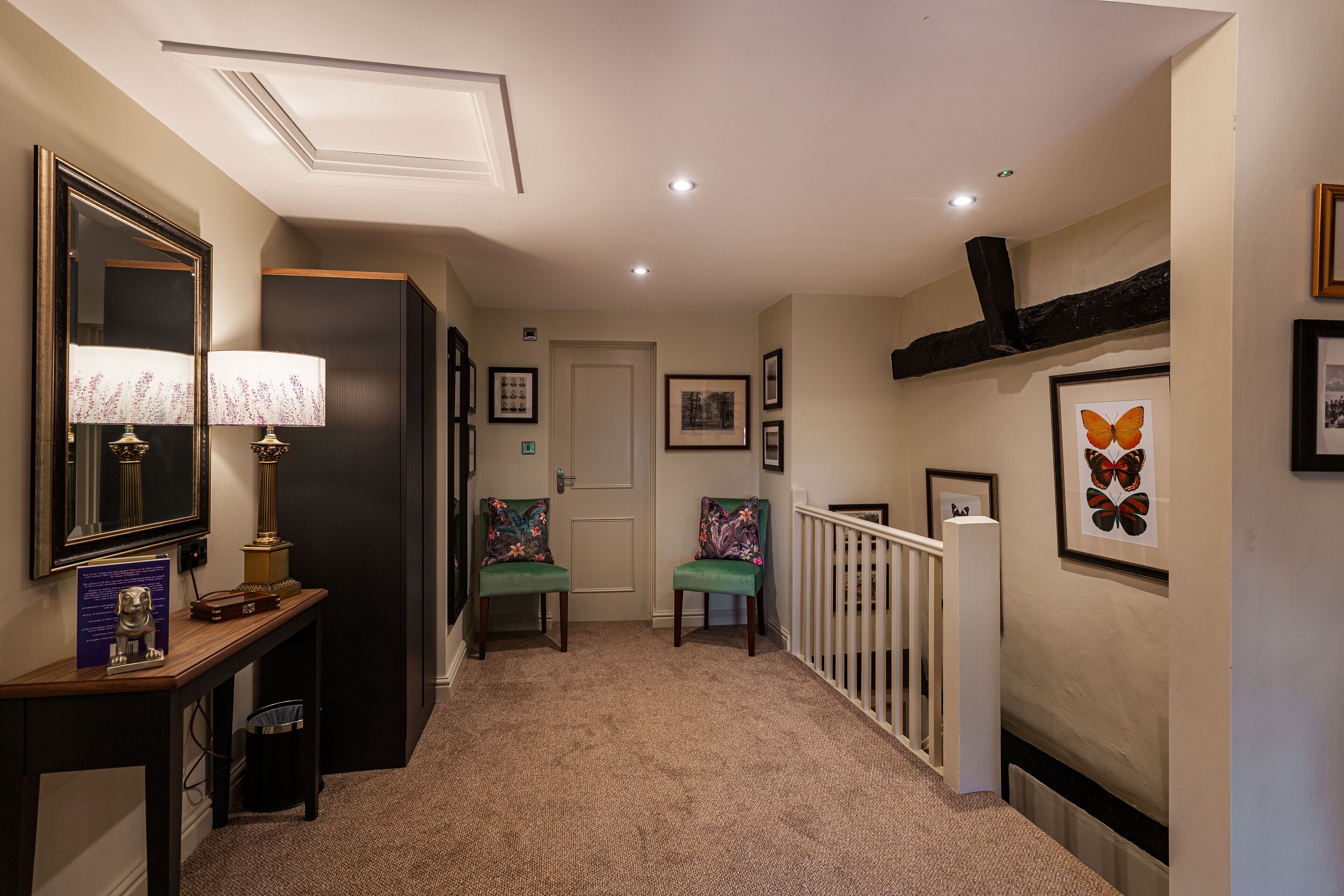 A hallway with a carpeted floor, a wooden console table with a lamp, two chairs, a white railing, and framed artwork on the walls.