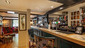 A bar area with a wooden counter, bar stools, and a variety of bottles and glasses. - The George Hotel, Dorchester-on-Thames (Wallingford)