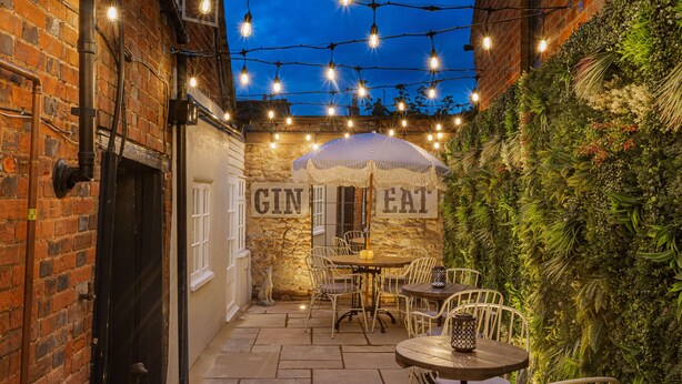 An outdoor patio with stone flooring, white metal chairs, a round table, and a hanging string of lights. - The George Hotel, Dorchester-on-Thames (Wallingford)