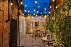 An outdoor patio with stone flooring, white metal chairs, a round table, and a hanging string of lights. - The George Hotel, Dorchester-on-Thames (Wallingford)