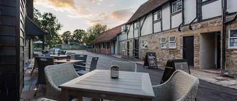 An outdoor seating area with wicker chairs and tables, a stone building with a tiled roof, and a clear sky.