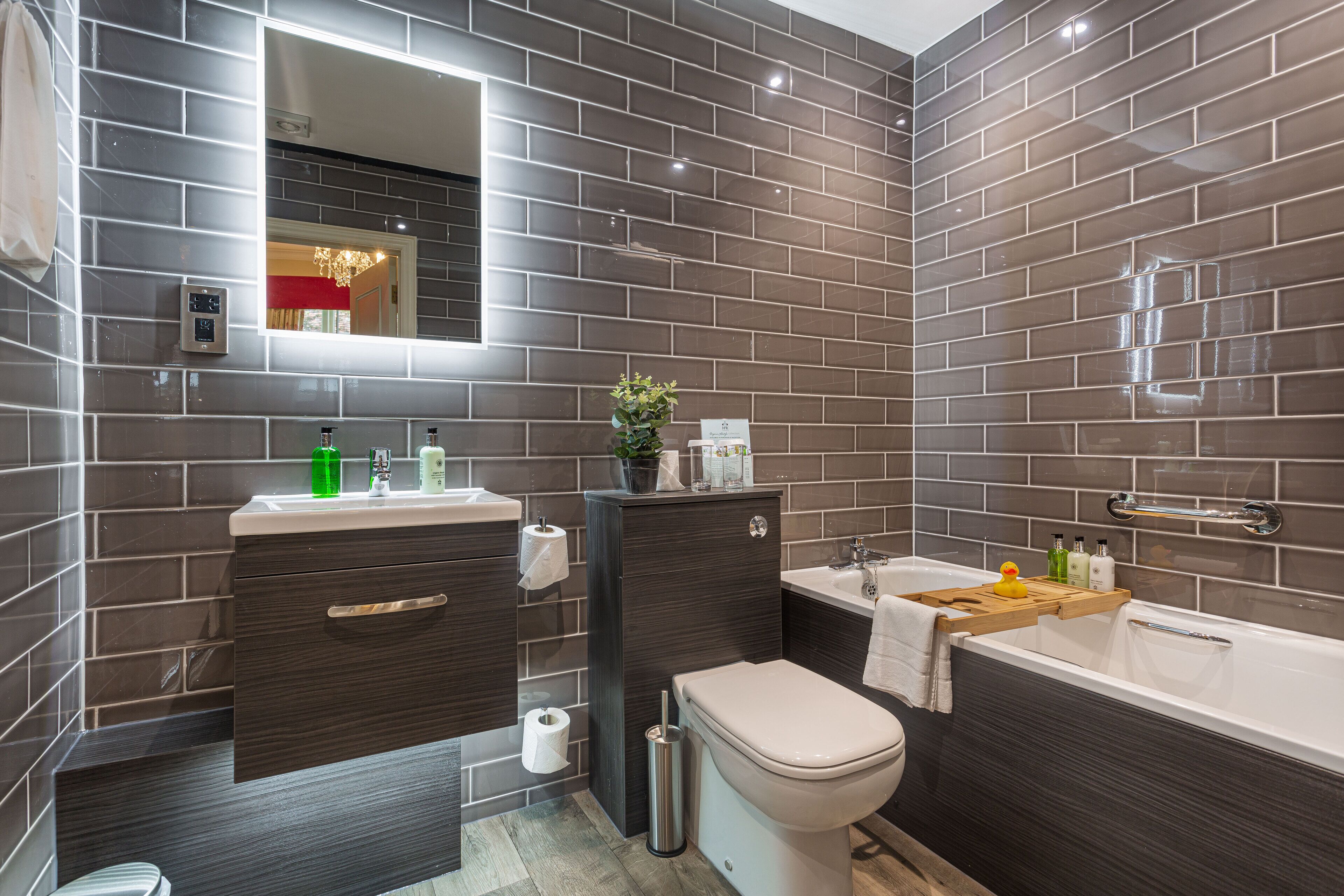 A modern bathroom with dark tiled walls, a white toilet, a sink with a mirror, and a bathtub with a wooden tray.