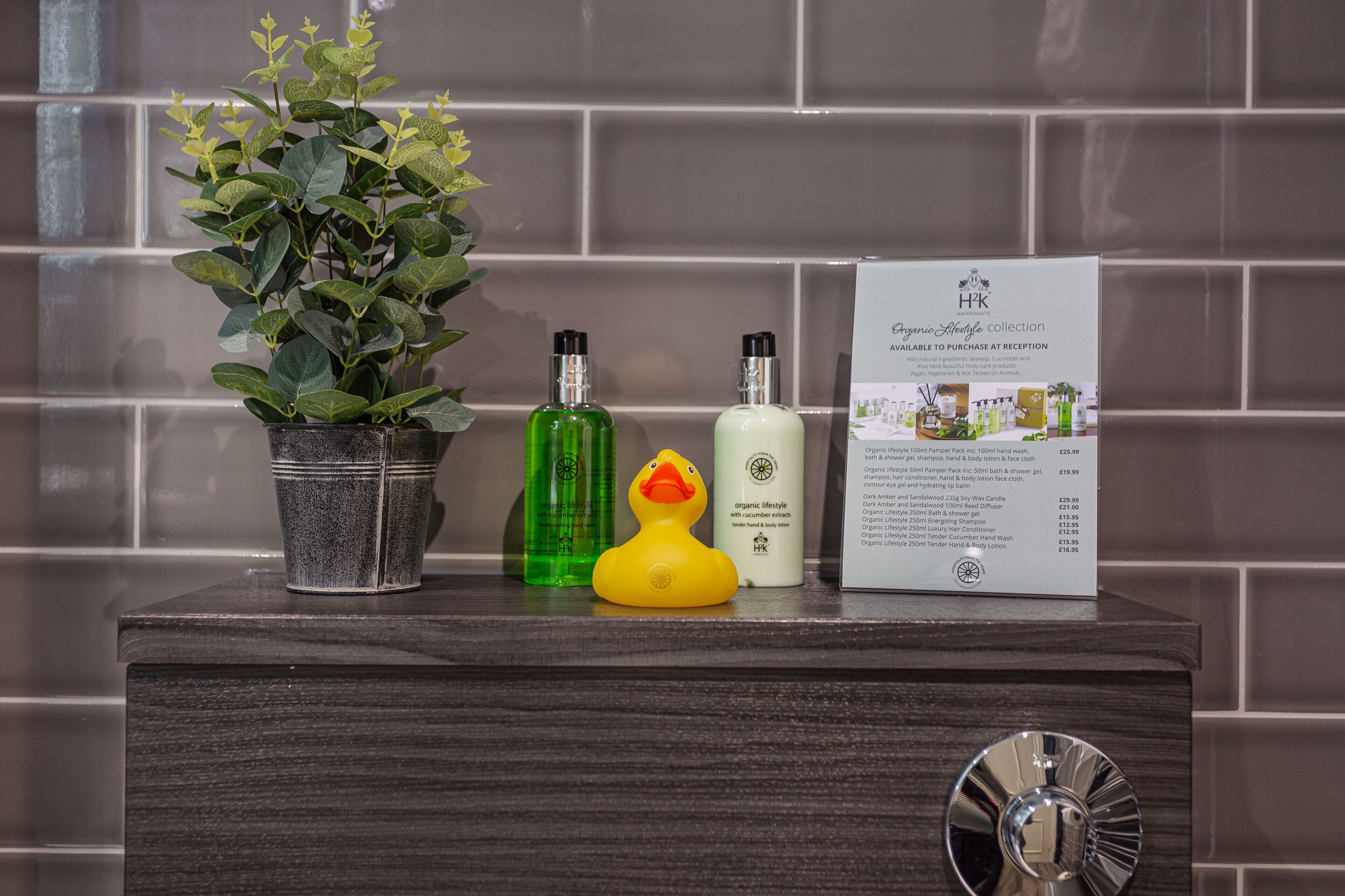 Un lavamanos de baño con una planta en maceta, dos botellas de productos orgánicos, un patito de goma y un folleto de productos.