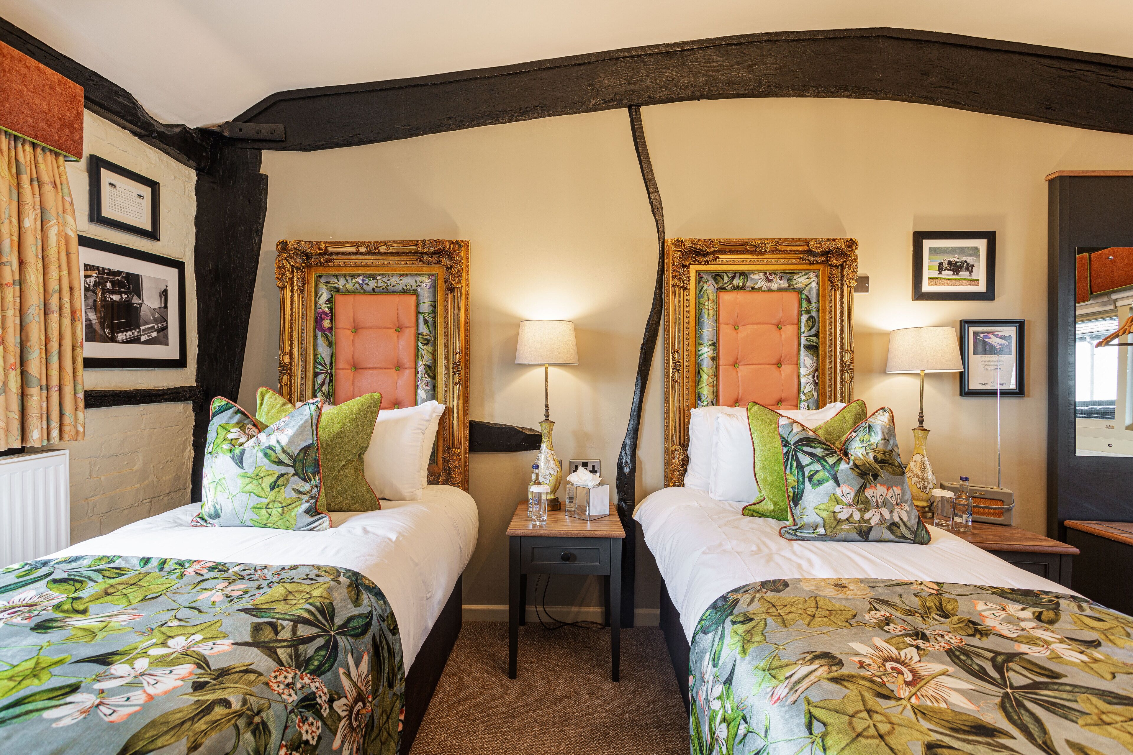 A hotel room with two beds, floral bedspreads, a nightstand with a lamp, and framed pictures on the walls.