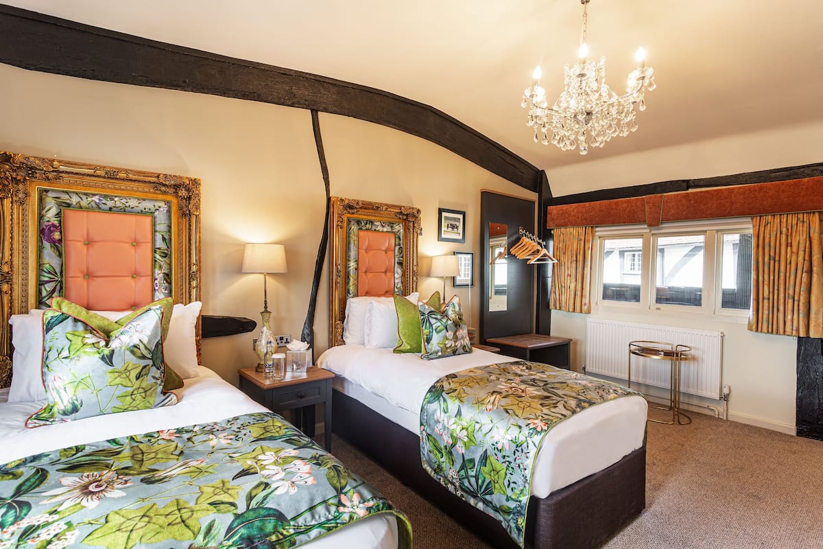 A hotel room with two beds, a chandelier, a window with curtains, and a radiator.