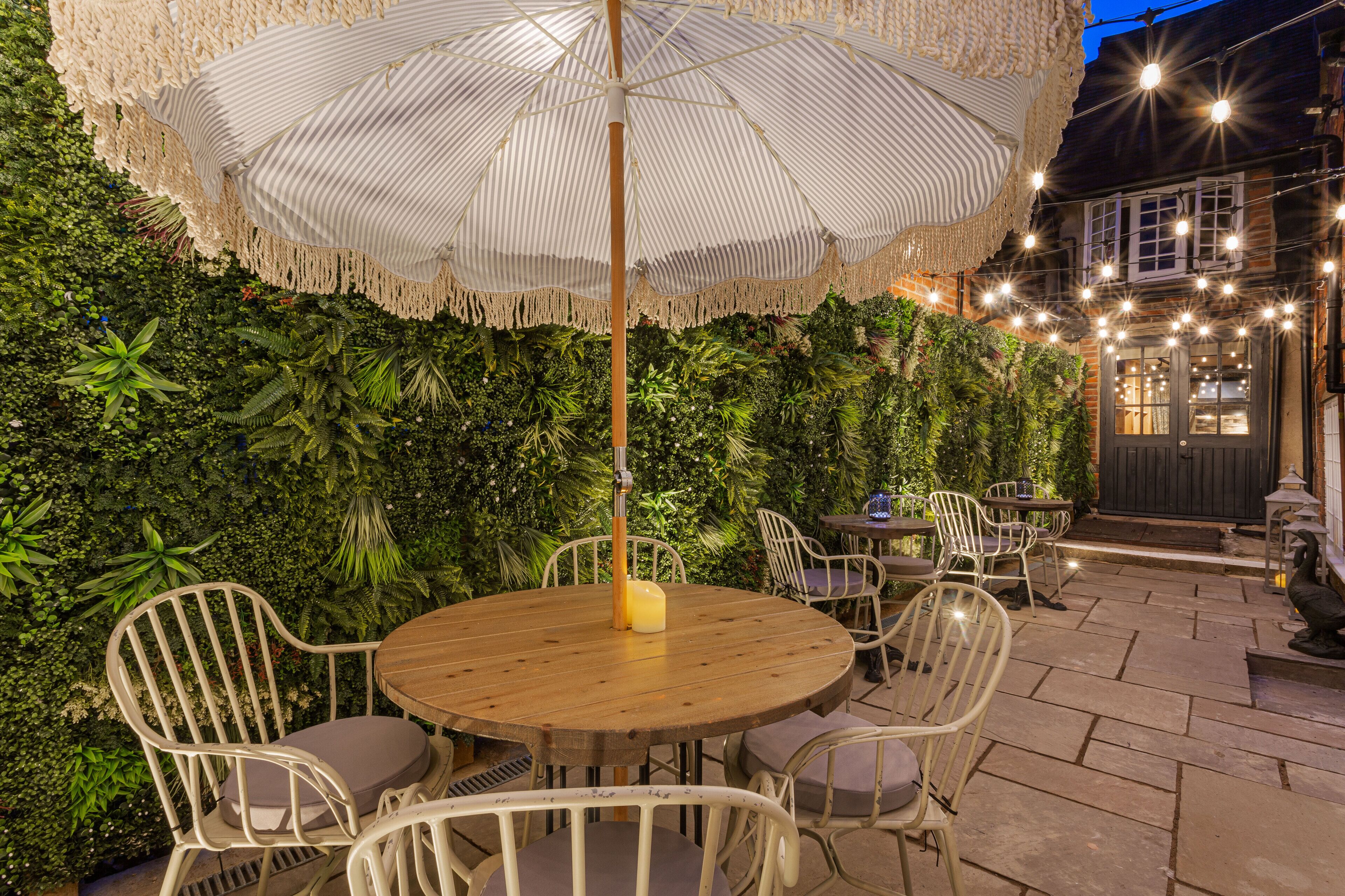 An outdoor patio with round wooden tables and white chairs, surrounded by lush greenery and string lights.