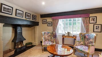 Une pièce avec une cheminée, deux fauteuils à motifs floraux, une table ronde en bois et des tableaux encadrés accrochés au mur.