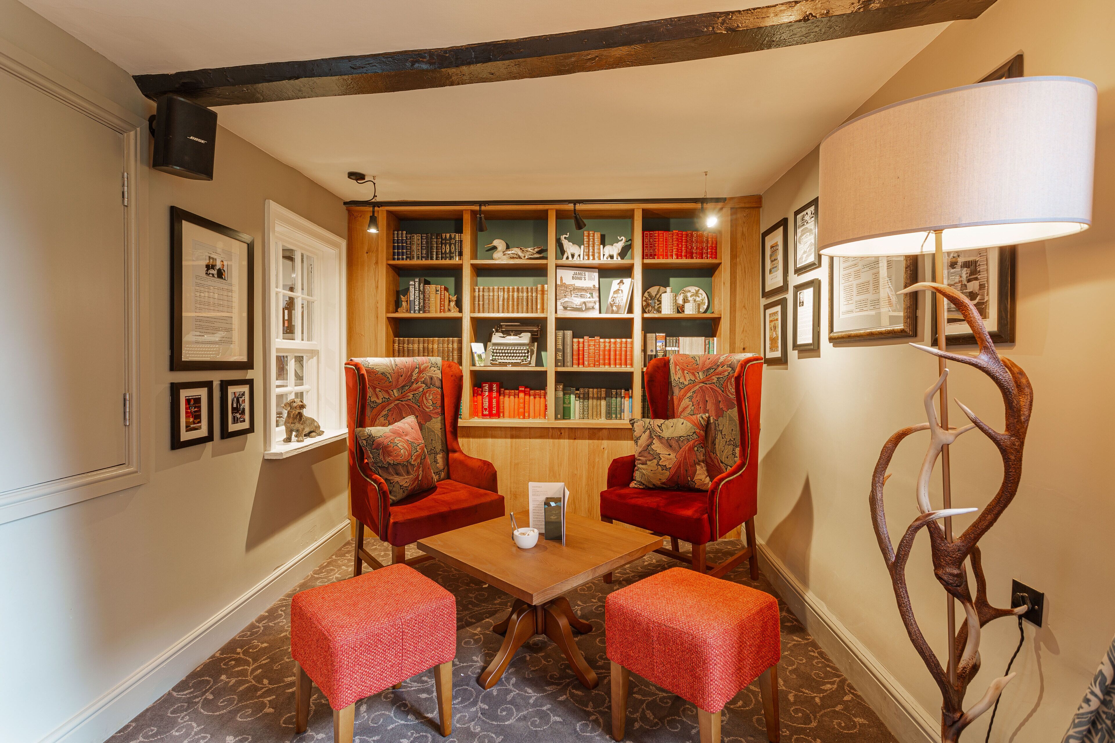 Un rincón acogedor para leer con dos sillones rojos, una mesa de madera y una estantería llena de libros y adornos.