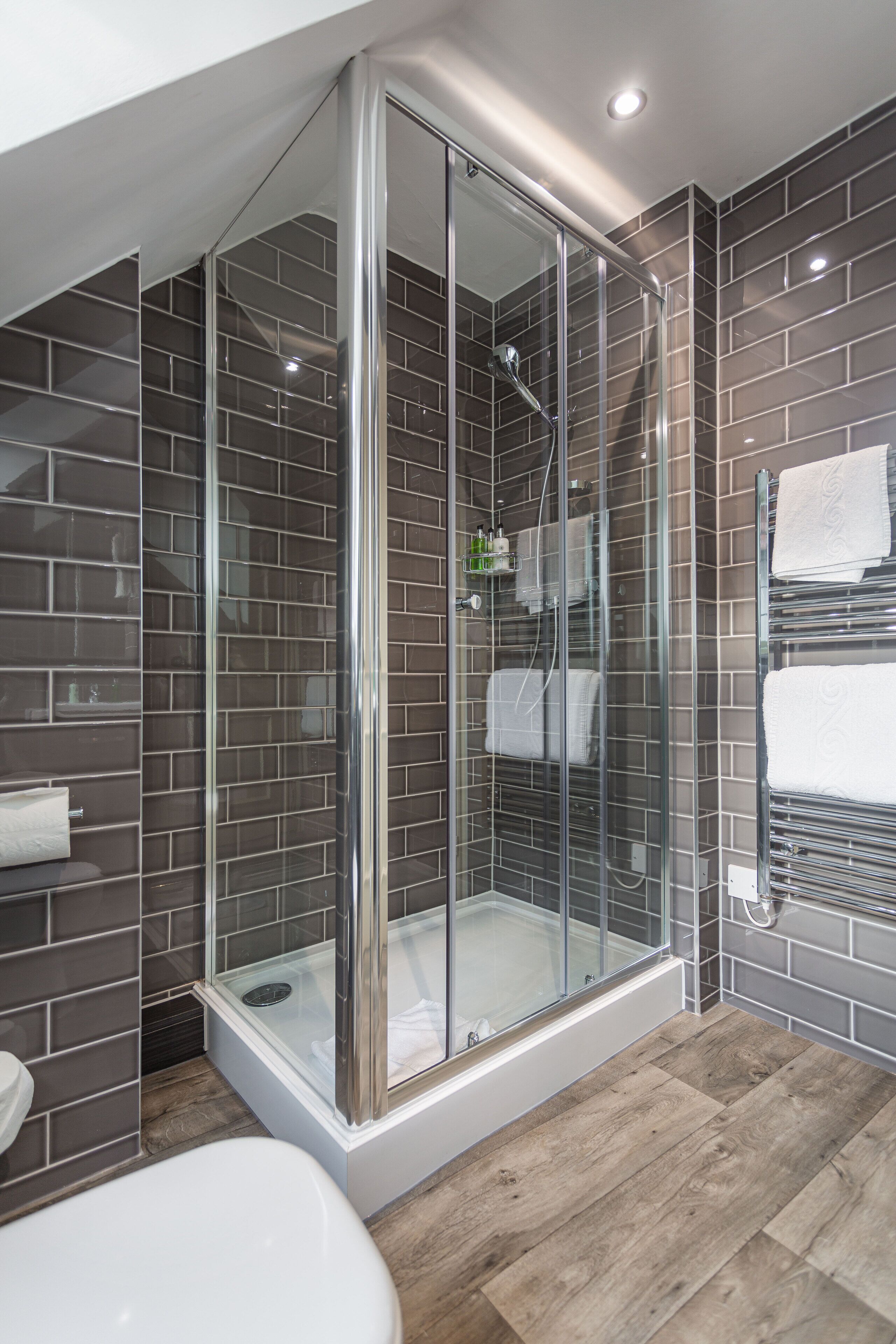 Un baño moderno con ducha cubierta por vidrio, paredes con azulejos oscuros y suelo de madera.
