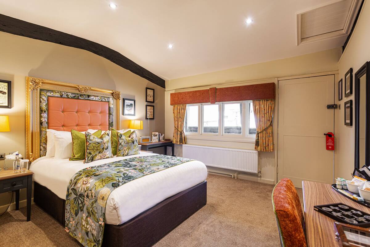 A hotel room with a large bed, a desk, a chair, a window with curtains, and a radiator.