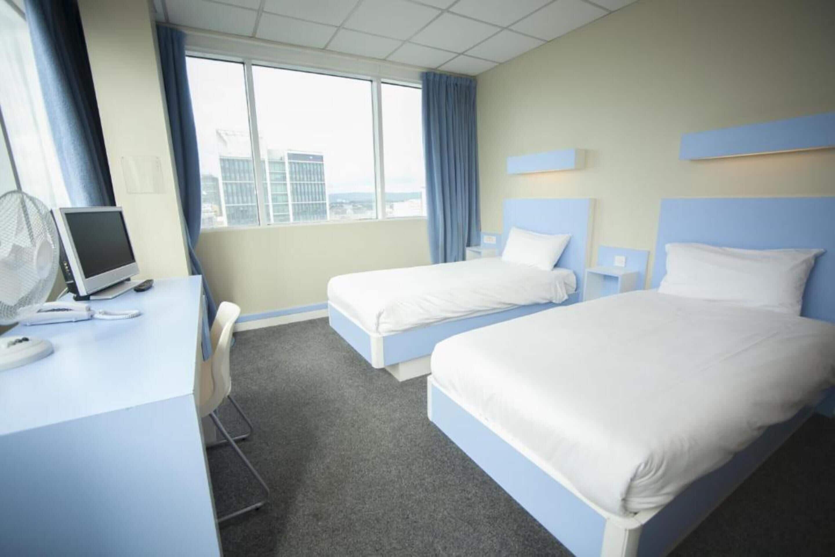 A hotel room with two beds, a desk with a computer, and a window with a view of buildings.