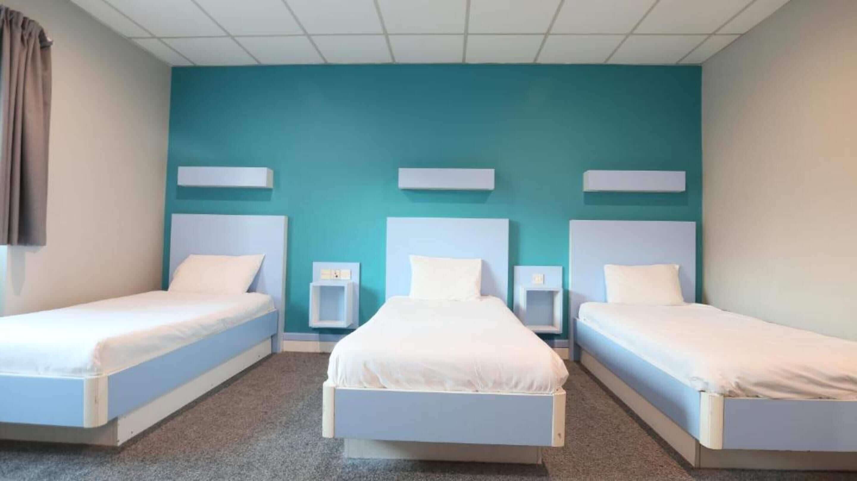 A hotel room with three single beds, each with a white headboard and matching nightstands. The walls are painted teal, and there is a window with curtains on the left side.