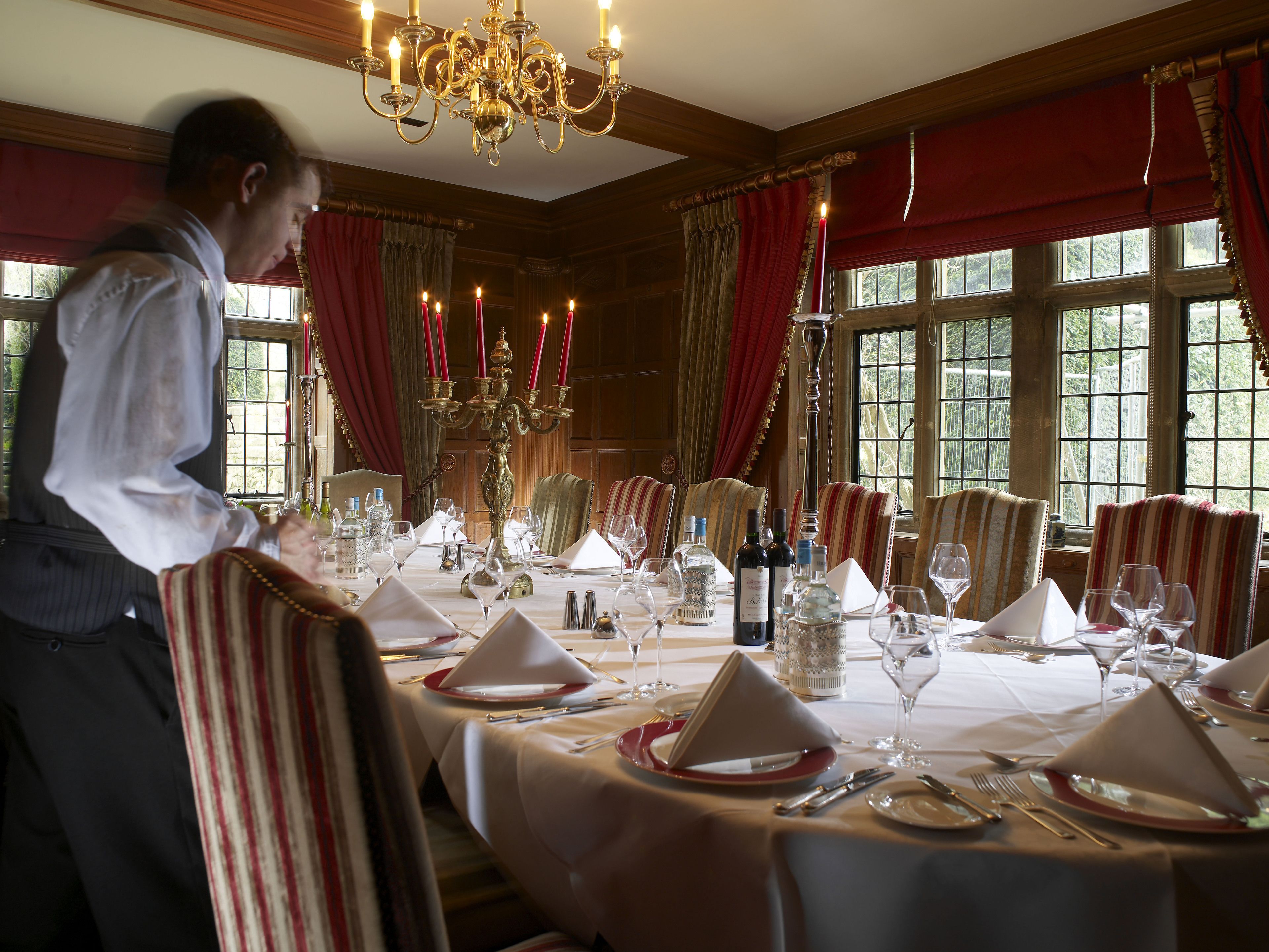 A formal dining room with a long table set for a meal, featuring red and white striped chairs, a chandelier, and large windows with curtains.