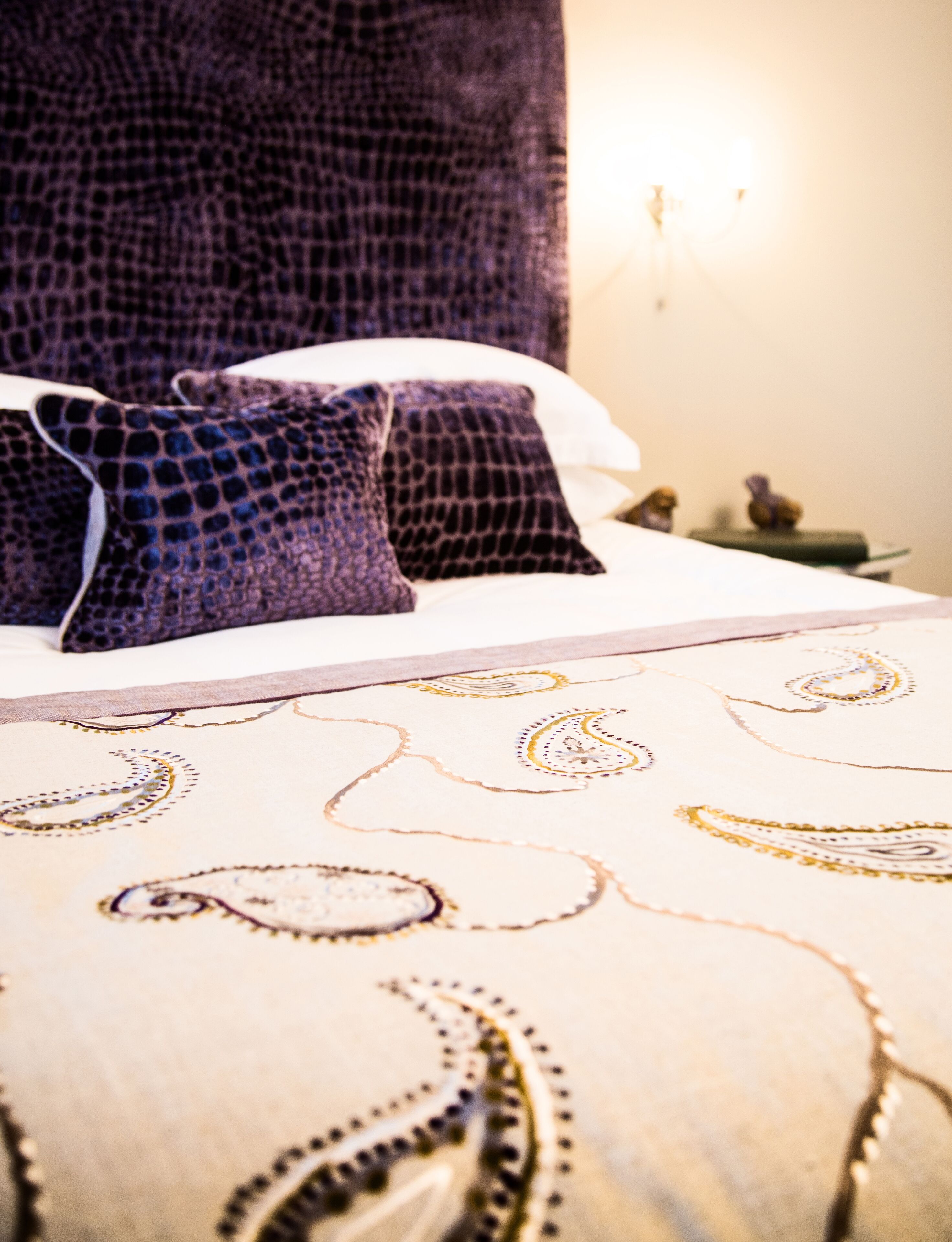 Un lit avec une couvre-lit à motifs et deux oreillers, une table de chevet avec une lampe et un mur muni d’un luminaire.