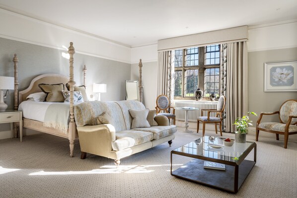 A bedroom with a four-poster bed, a sofa, a coffee table, and a view of the garden through a large window.