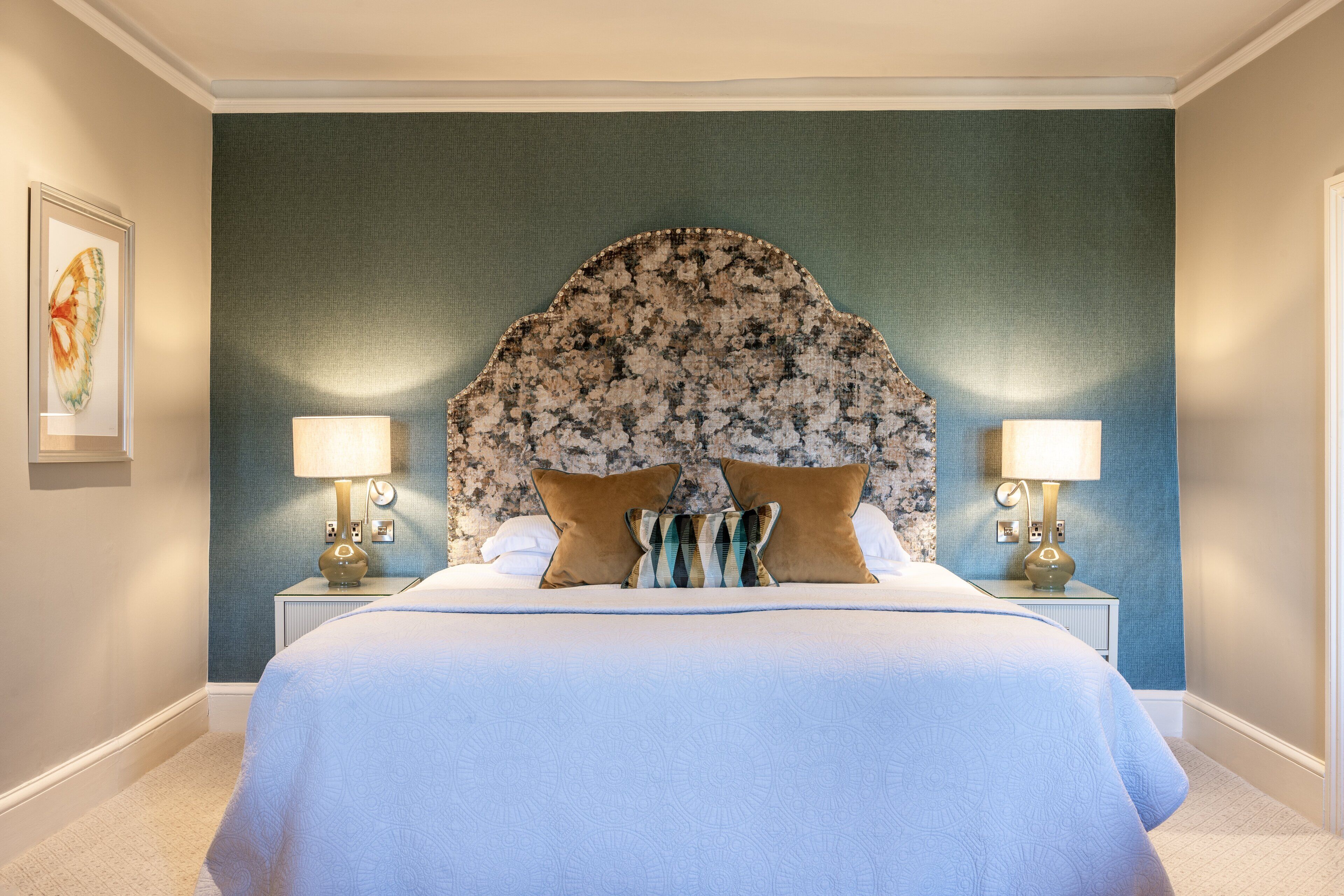 A bedroom with a large bed, two bedside lamps, a floral headboard, and a framed picture on the wall.